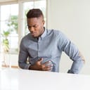 man with a hand on his stomach expressing pain due to indigestion, seated at a white table