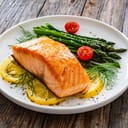 Close-up of a served fried salmon steak with green asparagus, fresh cherry tomato, and three slices of lemon.