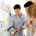 A group of people standing next to a board with architectural drawing looking at a tablet displaying a house project