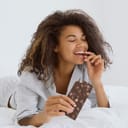 Dessert food and guilty pleasure concept. Happy woman eating chocolate with nuts while she lying in bed, wearing in pajamas. Smiling female biting candy bar, enjoying taste, spending morning at home
