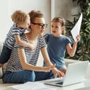 An exhausted young mom trying to focus on her laptop and paperwork on the couch with her small children.