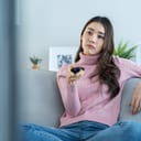 young woman watching a movie on TV with a bored expression on her face, sitting on a sofa at home