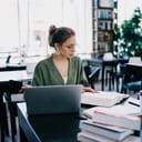 Young woman in glasses and elegant clothes sitting at desk among printed literature and laptop while reading book in modern library

