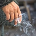 A person's hand holding a half-burnt cigarette with visible smoke coming out of it