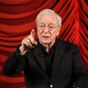 Elderly Michael Caine in black suit pointing his finger towards the audience