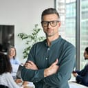 Smiling confident mature businessman leader looking at camera standing in office at team meeting. Male corporate leader ceo executive manager wearing glasses posing for business portrait arms folded.