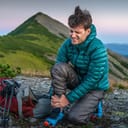 Young male traveler, with a backpack next to him, grimaces in pain and clutching a sprained ankle.