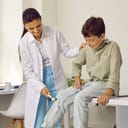 A female doctor using a hammer to check knee reflex of school boy sitting on medical couch in exam room