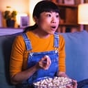 Asian woman at home late at night, eating popcorn and watching TV with a surprised face expression