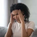 young woman sitting on a couch, frowning and touching her temples, experiencing discomfort from a strong headache