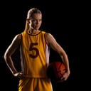 A young woman in a basketball uniform, holding the ball in one hand and standing against a black background.