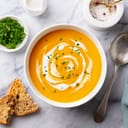 A bowlful of pumpkin soup next to freshly chopped green leaves, salt and red pepper mixed together, cream, couple of slices of bread and a spoon on the table
