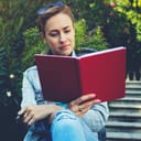 Young beautiful female tourist is reading interesting book during her rest after walking in the fresh air, charming woman student is preparing to the study at University while is sitting on a campus