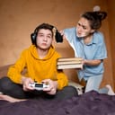 A mother lifting headphones from her son's head and giving him books while he is playing video games