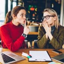A woman placing a finger over her mouth in order to tell the other not to say anything in an office