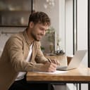 A smiling man sitting at a desk and writing on a piece of paper while using his laptop