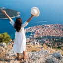 Girl with hands spread wide open standing on a hill looking down at Dubrovnik's old town