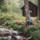 man sitting in front of a wooden cabin in the forest by a river