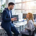 young businessman lounging on a desk and talking with a female colleague who is seated at her desk in the office