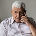 Serious concerned old grandpa talking on mobile phone, calling for emergency, ambulance. Senior 80s man speaking on cellphone, sitting on couch, at home, contact family on distance