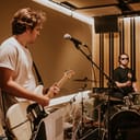 Two men playing a guitar and drums in a recording studio