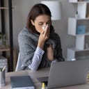 Sick girl wrapped in a scarf and blowing her nose, sitting in front of her laptop