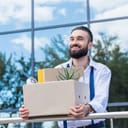 A happy young businessman standing outside an office building, holding a cardboard box with his office items after resigning.