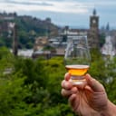 Hand holding glass of single malt scotch whisky and view from Calton hill to park and old parts of Edinburgh city in rainy summer day, Scotland, UK