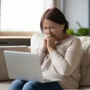 Sad mature woman looking at laptop screen and crying, sitting on couch at home