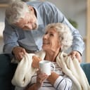 Affectionate elderly man covering shoulders of smiling middle aged hoary wife with plaid, showing care to beloved woman at home. Happy old mature retired family couple enjoying cozy weekend sweet time
