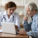 Confident doctor consulting mature patient at appointment, explaining, showing medical checkup results on laptop screen, serious young woman physician giving recommendations at meeting