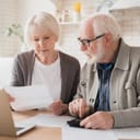 An elderly couple sitting at table in front of a laptop with a calculator looking at a piece of paper