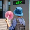A woman using a hand fan with a street thermometer showing 48 degrees Celsius