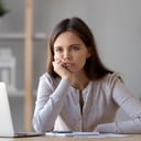 Bored woman sitting at table with laptop, pen and notepad in front of her