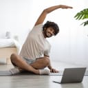 A young man sitting on a floormat in front of a laptop exercising yoga
