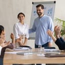 A smiling male leader praises his happy millennial Arab colleague during a group meeting, with diverse coworkers applauding.