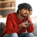 A bored young man sitting on a couch at home, watching TV, resting his head on his hand and flipping through channels with a remote control.