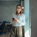 Businesswoman in casual attire standing in the office and smiling with a mobile phone in hand