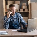 male salesperson referencing documents while confirming orders with a customer on the phone in a warehouse with laptop in front of him