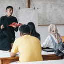 High school students attentively listening as a young male teacher explains something at the front of the classroom.