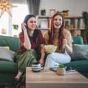 two happy sisters sitting on a sofa enjoying a movie or TV series at home and eating popcorn