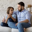 Serious father listen to his pre-teen little son talking seated on sofa at home, speaking spend time together at home. Cute boy share problems, ask advice to dad. Communication, care and trust concept