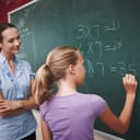 A teacher standing next to a girl who is doing math on a blackboard