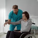Male nurse assisting a young paralyzed man in a wheelchair with arm exercises in his room at a rehabilitation center