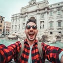 A selfie of a young man wearing glasses in a flannel shirt in front of the Trevi Fountain