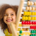 A Black student in a yellow dress laughs brightly behind a colored abacus in an elementary school classroom