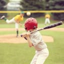 kid in safety gear swinging a bat on a baseball field with other players in the background