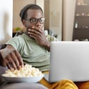 Man with shocked expression on his face eats popcorn while watching a movie on a laptop.