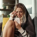 A young woman wiping her tears with a paper tissue while looking at the phone covered with a blanket