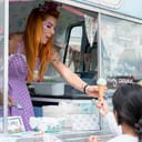 A woman with facepaint in an ice-cream truck giving an ice-cream to a girl